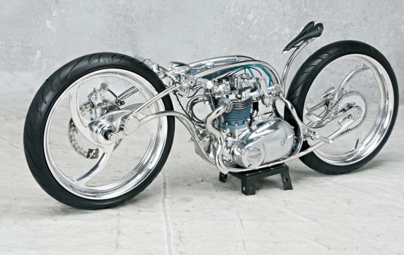 «Серебро» чемпионата, звание «метрический чемпион» и приз Partner Pick Of Excellence от городского совета Стерджиса – такой урожай наград собрал американец Крис Кром (Kris Krome Customs) с помощью этого странного, с позволения сказать, мотоцикла под названием Re-flex-tion. 650-кубовая «двойка» и коробка передач – от Triumph T120 Bonneville. Рама-риджид – из нержавеющей стали (здесь вообще «ни капли» хрома – сплошь полированная нержавейка). Передняя подвеска – типа Elastamerick. Стоп! А вилка где? И вообще – как «это» управляется? Ручки-то к раме приварены! А вот тут-то и кроется главная фишка. Если присмотреться, можно увидеть шарнирные соединения в двух точках – перед двигателем и под сиденьем. Условная прямая между ними – по сути и есть рулевая колонка. То есть чтобы повернуть, надо «завалить» всю верхнюю часть рамы. Рейк получается зверский – 75o, но мотоцикл с грехом пополам рулится. Колеса – 23-дюймовые. Шины – «стотридцатки» Avon. Задний тормоз конструкцией не предусмотрен, передний – Jaybrake.